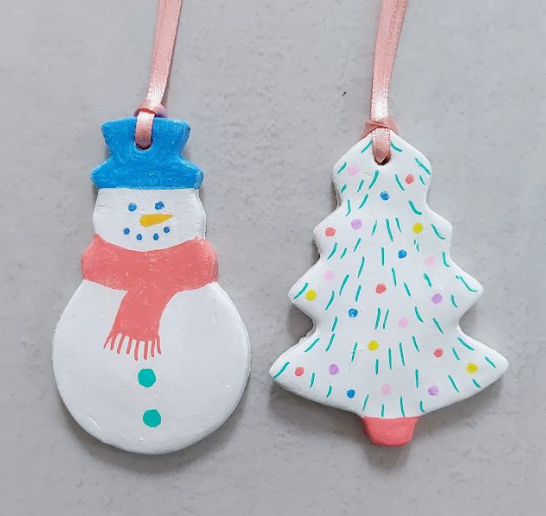 Two festive air dry clay crafts shaped into hanging Christmas ornaments featuring a snowman wearing a blue hat and pink scarf alongside a decorated holiday tree with colorful dots.