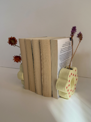 Two creative air dry clay crafts shaped into flower-patterned bookends supporting a stack of books, featuring yellow ceramic-look pieces painted with red crosses and holding dried flowers.
