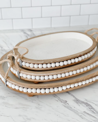 A stack of three oval wooden trays decorated with white bead detailing along the rims and rustic rope handles, displayed against a white tile backsplash.
