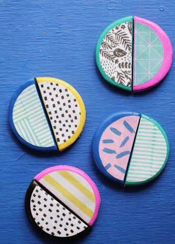 An overhead shot of four circular air dry clay crafts styled as decorative coasters with split patterned halves, featuring geometric prints, polka dots, stripes, and brushstrokes on a bright blue background