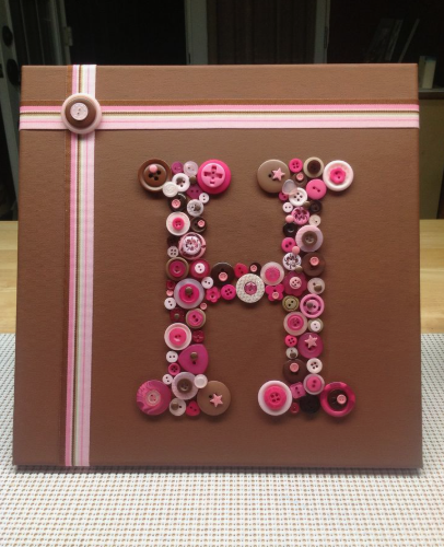 30 Creative DIY Button Crafts to Turn Spare Buttons Into Beautiful Decor A brown canvas wall art project featuring a monogram letter H beautifully outlined with an assortment of pink and brown buttons, accented with striped ribbons, showcasing DIY button crafts.