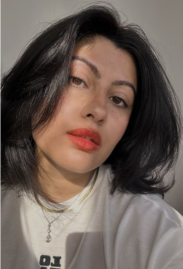 A close-up selfie of a woman with dark shoulder-length layered hair, wearing a white t-shirt and vibrant coral-orange lipstick.