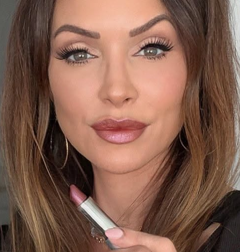Spring 2026 lipstick trends featuring a woman with long brown hair holding a silver lipstick tube while wearing a glossy neutral pink shade with defined lip liner.