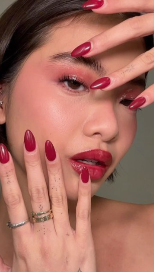 Spring 2026 lipstick trends featuring a glossy cranberry-red lipstick paired with matching almond-shaped red nails and coordinated monochromatic eye makeup.