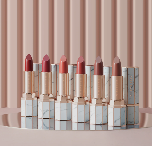 Spring 2026 lipstick trends featuring a line-up of six luxury lipsticks in octagonal marble and rose-gold cases, displaying a spectrum of shades from red to nude on a reflective platform.