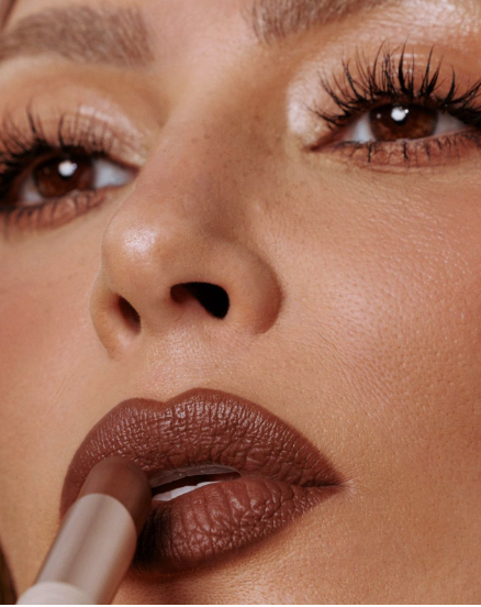 Spring 2026 lipstick trends featuring a close-up macro view of a person applying rich brown matte lipstick to their lips with a white cosmetic tube.