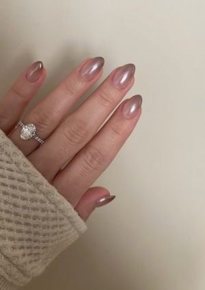 Short almond-shaped neutral taupe chrome nails with a pearlescent glazed finish, paired with a diamond ring and beige sweater.