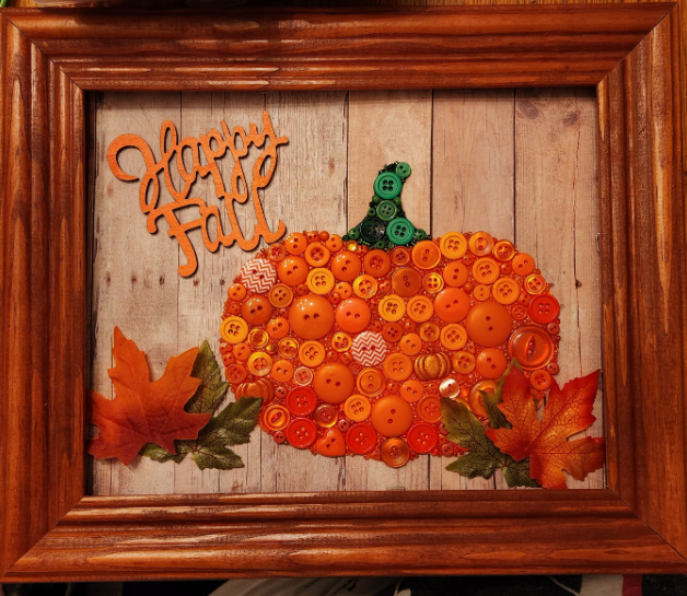 30 Creative DIY Button Crafts to Turn Spare Buttons Into Beautiful Decor A framed autumn-themed wall art piece featuring a pumpkin made of orange and yellow buttons with a green button stem, fabric autumn leaves, and the phrase "Happy Fall", highlighting DIY button crafts.