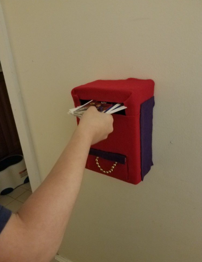 20 Recycled Household Item Crafts for a Creative Home A wall-mounted mailbox created as a recycled household item craft from a cardboard box covered in red and purple fabric, with a hand sliding mail into the front slot.