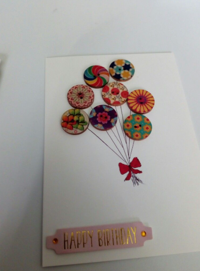 30 Creative DIY Button Crafts to Turn Spare Buttons Into Beautiful Decor A handmade birthday greeting card featuring a cluster of colorful patterned wooden buttons tied with drawn string lines to a red bow, with the gold-foiled text "HAPPY BIRTHDAY" at the bottom, highlighting DIY button crafts.