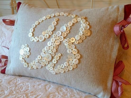 30 Creative DIY Button Crafts to Turn Spare Buttons Into Beautiful Decor A rustic beige linen throw pillow accented with a monogram letter formed from white buttons and tied with red and white checkered ribbon bows at the corners, showcasing DIY button crafts.