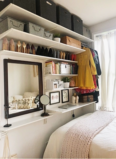 Small bedroom idea with no closet featuring extensive vertical wall shelving, a shoe display shelf, a hanging clothing rack next to the bed, and a wall-mounted mirror and desk workspace.