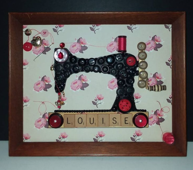30 Creative DIY Button Crafts to Turn Spare Buttons Into Beautiful Decor A framed mixed-media wall art piece featuring a vintage sewing machine silhouette meticulously crafted from black and red buttons, set against a floral background with Scrabble tiles spelling the name "LOUISE" at the base, highlighting DIY button crafts.