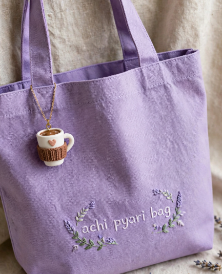 DIY clay bag charms featuring a cute coffee mug with a cozy knitted sleeve attached to a light purple canvas tote bag embroidered with "achi pyari bag" and floral details.