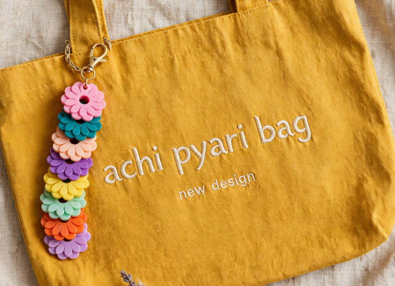 DIY clay bag charms featuring a vertical stack of colorful daisy-shaped charms attached to a mustard yellow canvas tote bag embroidered with "achi pyari bag new design".