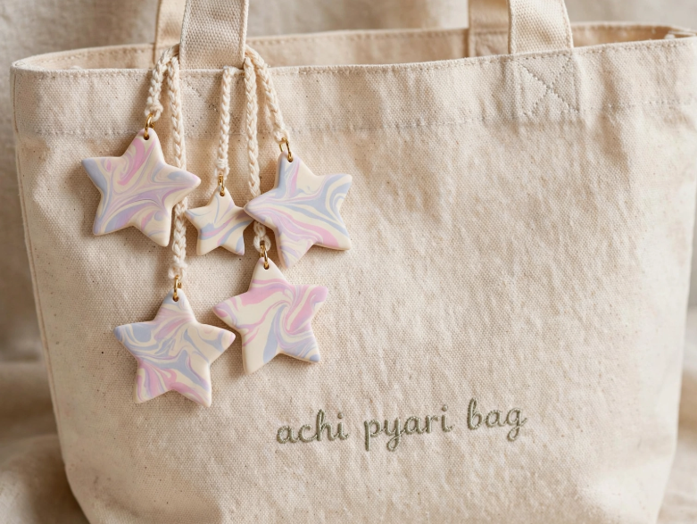 DIY clay bag charms featuring marbled pastel star-shaped charms hanging on braided cords attached to an off-white canvas tote bag embroidered with "achi pyari bag"
