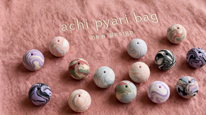 DIY clay bag charms featuring an array of colorful marbled spherical beads scattered on a pink fabric background with "achi pyari bag new design" embroidery.
