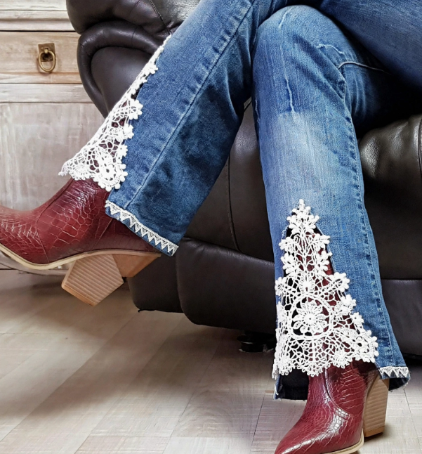 A person sitting on a dark leather couch wearing blue jeans styled with white crochet lace inserts at the cuffs and maroon boots, featured in 15 Crochet Upcycle Ideas.