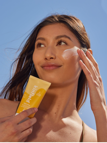 Model applying Kinfield sunscreen to her cheek under a blue sky, highlighting Fall 2026 Beauty Trends.