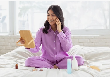 Smiling woman in purple pajamas sitting on a bed looking into a handheld mirror while trying out skincare items aligned with Fall 2026 Beauty Trends.