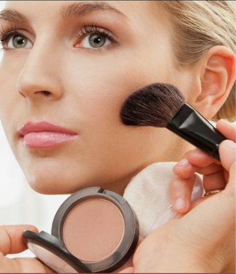 Low Maintenance Beauty Routine: A Simple 5-, 10-, and 15-Minute System A close-up of a woman applying powder blush with a makeup brush as part of a low maintenance beauty routine, holding a compact mirror with a pressed powder pan.