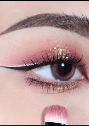 Low Maintenance Beauty Routine: A Simple 5-, 10-, and 15-Minute System A close-up shot of a detailed eye makeup look featuring pink eyeshadow, gold glitter, sharp winged eyeliner, and a makeup brush gently dusting powder beneath the eye as part of a low maintenance beauty routine.