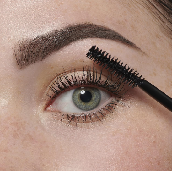 Low Maintenance Beauty Routine: A Simple 5-, 10-, and 15-Minute System A close-up shot of a green eye with detailed eyelashes as a mascara wand applies mascara, paired with a neatly filled brow as part of a low maintenance beauty routine.