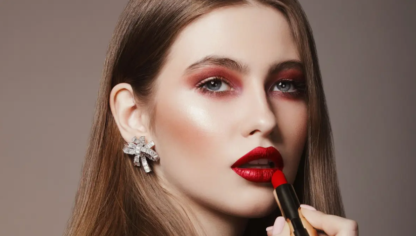 Low Maintenance Beauty Routine: A Simple 5-, 10-, and 15-Minute System A close-up of a woman with red eyeshadow and statement earrings as she applies red lipstick, representing a polished look that complements a low maintenance beauty routine.