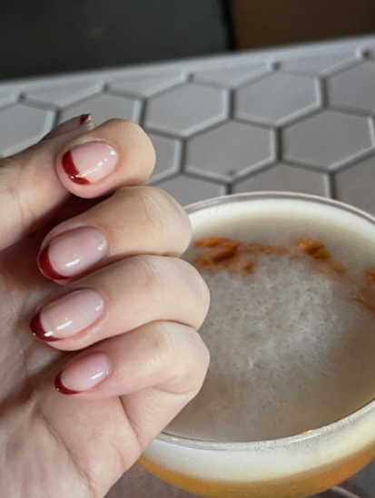 35 Short Nail Designs That Make Your Fingers Look Longer A close-up of short, rounded nails featuring a deep red French tip on a sheer base next to a cocktail, showcasing one of the short nail designs that make your fingers look longer.