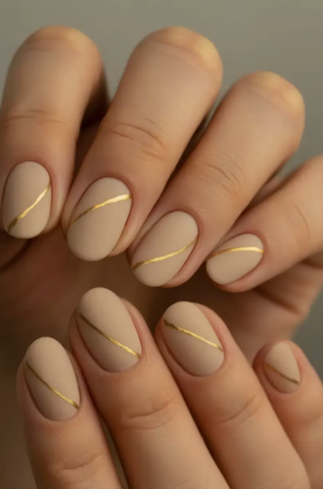 35 Short Nail Designs That Make Your Fingers Look Longer A close-up of short, oval-shaped matte nude nails featuring an elegant diagonal metallic gold line, serving as one of the short nail designs that make your fingers look longer.