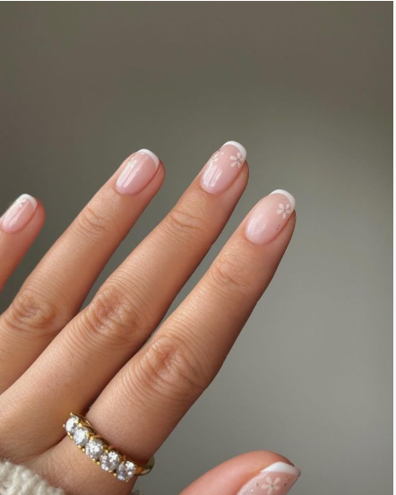 35 Short Nail Designs That Make Your Fingers Look Longer A close-up of short, rounded nails featuring classic white French tips accented with tiny white floral details on a sheer base, serving as one of the short nail designs that make your fingers look longer.