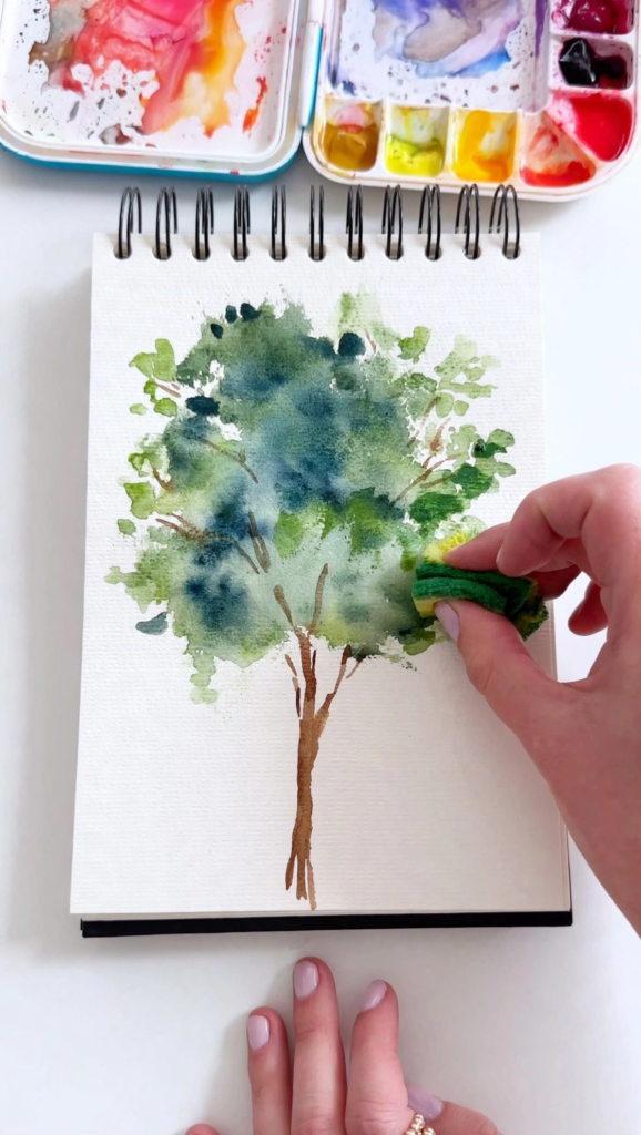 Hand dabbing a small sponge with green paint to create tree foliage in a watercolor sketchbook, showcasing analog crafts to do instead of scrolling.   
