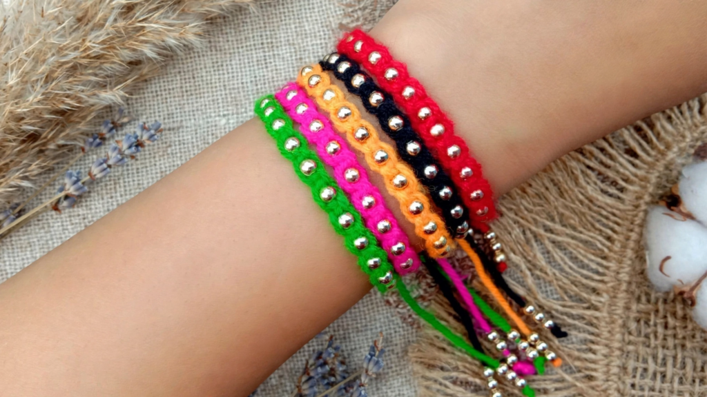 Hand wearing a stack of colorful braided yarn friendship bracelets with silver beads in green, pink, orange, black, and red, showcasing analog crafts to do instead of scrolling.   