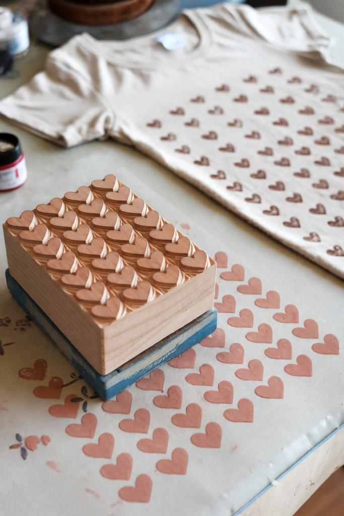 Carved wooden heart pattern stamp next to stamped fabric and a heart-printed t-shirt, showcasing analog crafts to do instead of scrolling.   