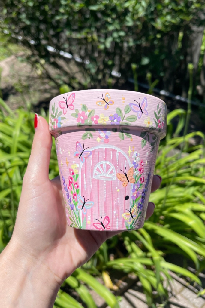 Handmade pastel pink painted terracotta flower pot with fairy door and butterflies, showing creative craft ideas for teens[cite: 23].