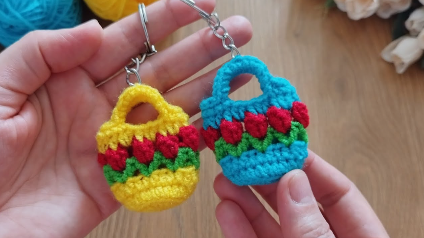 50+ Easy DIY Crafts to Sell and Make Extra Money Easy DIY crafts to sell: Two miniature crochet handbag keychains held in a person's hands, with the yellow one on the left and the bright blue one on the right, both featuring embroidered red tulip designs and top handles attached to metal key rings.