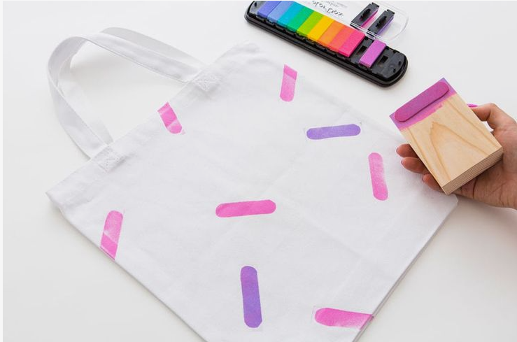 50+ Easy DIY Crafts to Sell and Make Extra Money Easy DIY crafts to sell: A white canvas tote bag being decorated with a wooden block stamp to print pink and purple dash patterns resembling sprinkles, with a rainbow ink pad set positioned in the upper right corner.