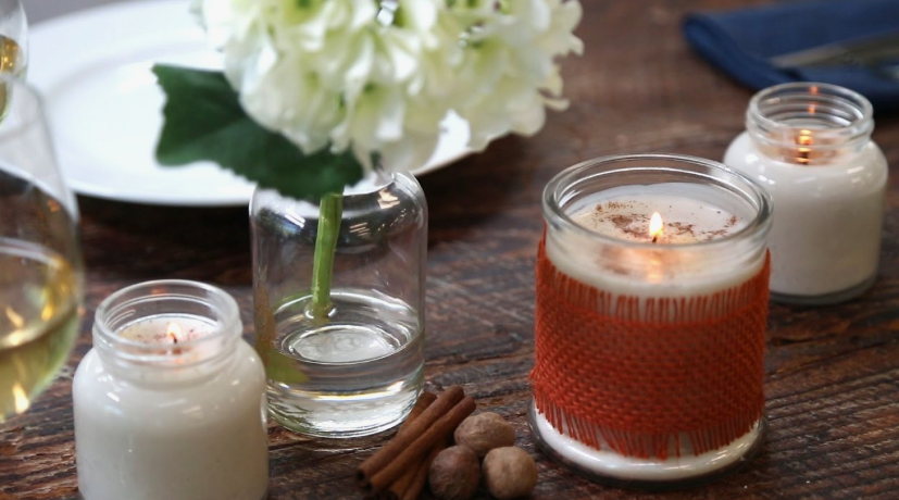 50+ Easy DIY Crafts to Sell and Make Extra Money Easy DIY crafts to sell: Homemade scented candles in small glass jars, including one wrapped in a textured orange yarn cozy and dusted with cinnamon, displayed on a rustic wooden table alongside cinnamon sticks, whole nutmeg, and a small vase with white flowers.