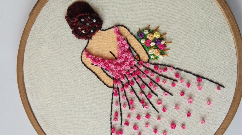 50+ Easy DIY Crafts to Sell and Make Extra Money Easy DIY crafts to sell: A hand-embroidered hoop art piece depicting a woman seen from behind, wearing a pink floral gown made of French knots and holding a colorful bouquet, set against an off-white fabric background.