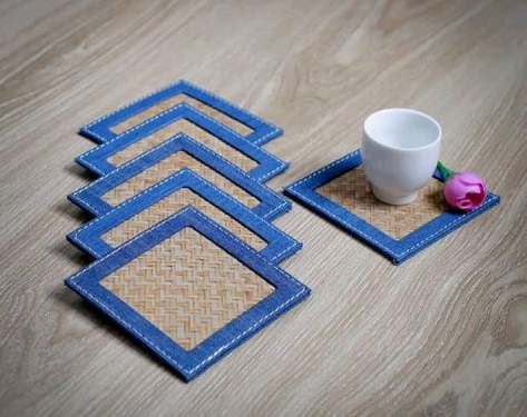 50+ Easy DIY Crafts to Sell and Make Extra Money Easy DIY crafts to sell: A set of square woven coasters edged in blue denim fabric with white stitching, featuring a stacked pile of four coasters on the left and a fifth coaster holding a small white teacup and a tiny pink flower on the right.
