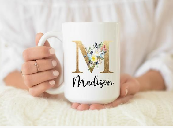 50+ Easy DIY Crafts to Sell and Make Extra Money Easy DIY crafts to sell: A white ceramic mug held by a person, featuring a large gold foil initial letter "M" decorated with blue and yellow floral accents and the name "Madison" written underneath in a black script font.