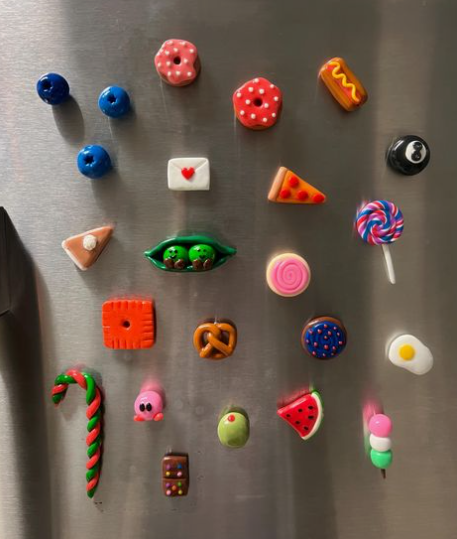 50+ Easy DIY Crafts to Sell and Make Extra Money Easy DIY crafts to sell: A collection of colorful miniature polymer clay food magnets attached to a metal surface, featuring items like donuts, pizza slices, a lollipop, a candy cane, a fried egg, and a pea pod.