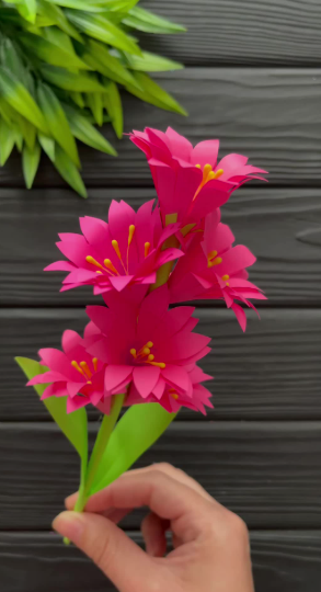 50+ Easy DIY Crafts to Sell and Make Extra Money Easy DIY crafts to sell: A handmade paper flower bouquet held in a hand, featuring vibrant pink blossoms with bright yellow stamen centers and green stems with leaves against a dark slatted background.