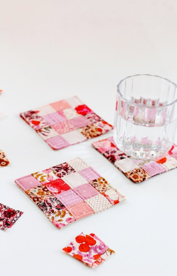 50+ Easy DIY Crafts to Sell and Make Extra Money Easy DIY crafts to sell: A set of handmade quilted fabric coasters featuring miniature patchwork squares in shades of pink, red, and white, with one coaster supporting a clear glass of water.