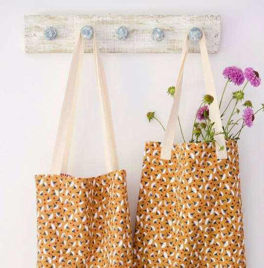 50+ Easy DIY Crafts to Sell and Make Extra Money Easy DIY crafts to sell: Two handmade mustard-yellow tote bags hanging from a rustic white wooden wall rack, featuring a repeating white and red leaf pattern, with one bag holding fresh purple flowers.