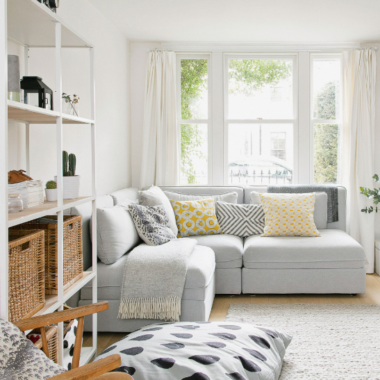Small Living Room Decor Ideas on a Budget: 25 Stylish Ways to Make It Look Expensive Bright and cozy small living room decor ideas on a budget featuring a light gray sectional sofa, tall white open shelving with woven storage baskets, yellow and patterned accent pillows, and floor-to-ceiling white curtains.