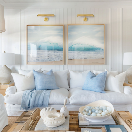 Small Living Room Decor Ideas on a Budget: 25 Stylish Ways to Make It Look Expensive Bright living room featuring small living room decor ideas on a budget with a white sofa, blue throw pillows and a matching throw blanket, twin ocean art pieces with picture lights, and a wooden coffee table filled with decorative bowls and stones.