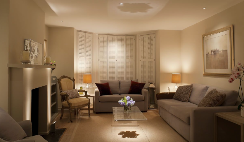 Small Living Room Decor Ideas on a Budget: 25 Stylish Ways to Make It Look Expensive Warmly lit living room featuring small living room decor ideas on a budget with a light gray sofa and loveseat, a glass coffee table, a fireplace with accent lighting, plantation shutters on bay windows, and framed wall art.