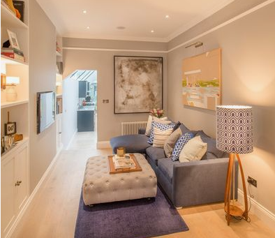Small Living Room Decor Ideas on a Budget: 25 Stylish Ways to Make It Look Expensive Compact living room featuring small living room decor ideas on a budget with a blue sectional sofa, a tufted light gray ottoman on a blue rug, built-in shelving, a wall-mounted TV, and a tripod floor lamp with a patterned shade.