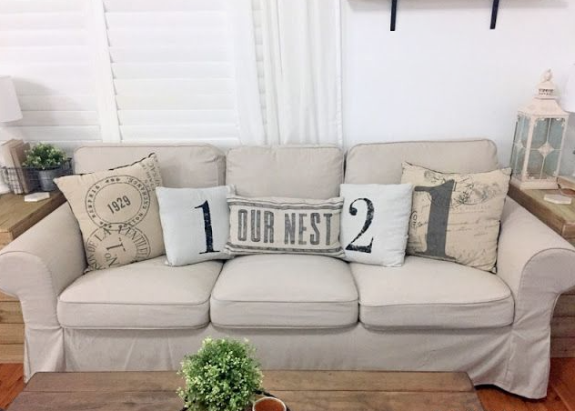 Small Living Room Decor Ideas on a Budget: 25 Stylish Ways to Make It Look Expensive Cozy living room featuring small living room decor ideas on a budget with a light beige slipcovered sofa adorned with number and typography accent pillows, wooden side tables, and a decorative lantern.
